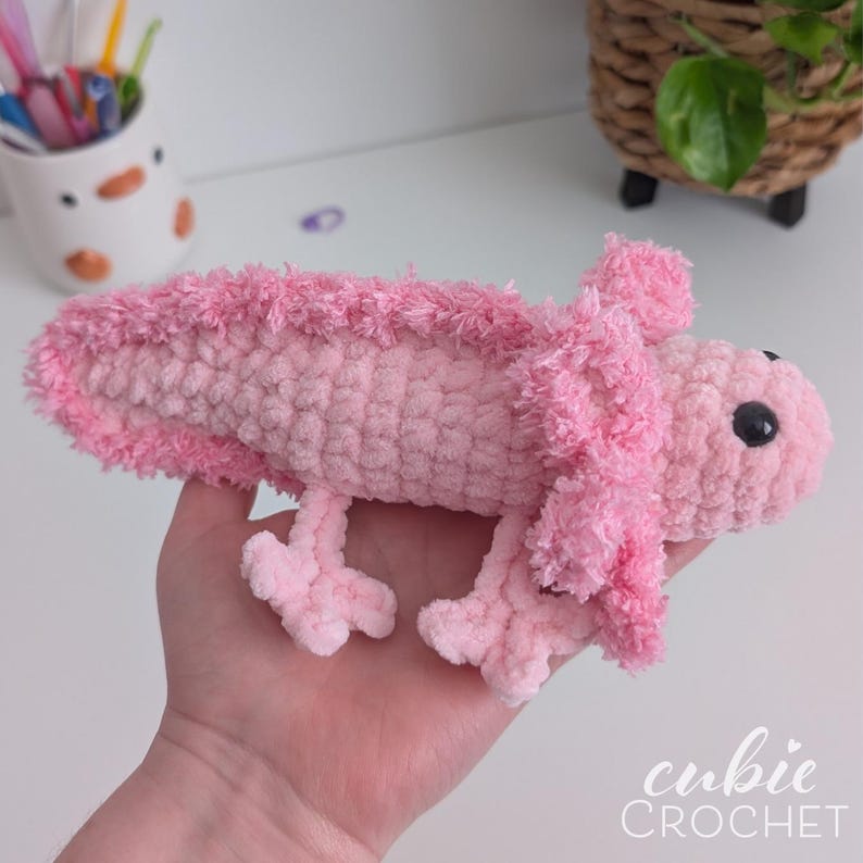 May include: A handmade pink crochet axolotl plush toy. The plush has a textured body, fluffy gills, and small black eyes. The axolotl is held in a hand, showcasing the soft texture and detail of the craft. The "cubie CROCHET" logo is visible.