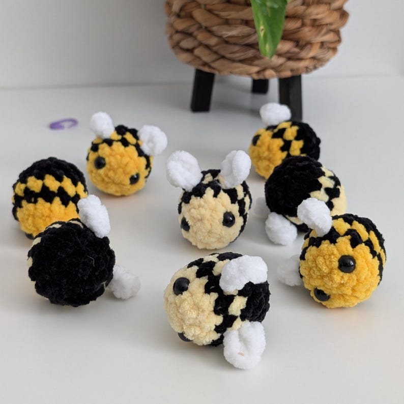 May include: A collection of handmade crochet bees in yellow and black, with white fuzzy wings and small black eyes. The bees are scattered on a white surface, with a woven basket in the background.