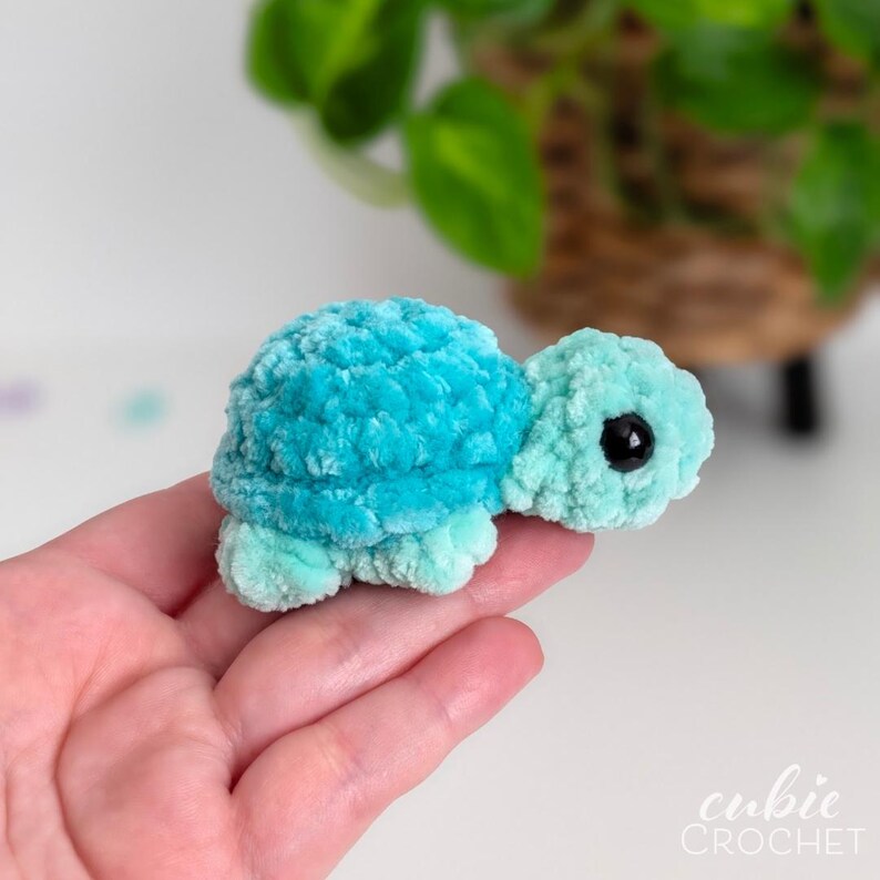 May include: A small, plush, crocheted turtle toy made from soft, fuzzy yarn. The turtle features a vibrant turquoise shell and a lighter mint green head and flippers. It has a single visible black plastic eye on the side of its head.
