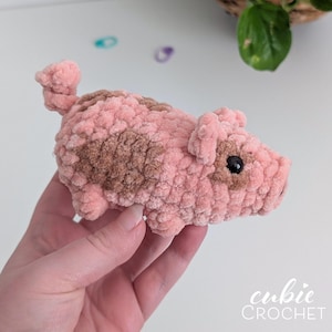 May include: A small, plush, crocheted pig toy made from soft, textured pink yarn with brown patches on its side and face. The pig features a curly tail, two small ears, and black bead eyes. The text 'cubie CROCHET' is visible in the bottom corner.