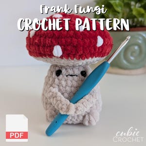 Puede incluir: Una figura de hongo de ganchillo con un gorro rojo y manchas blancas, sosteniendo un ganchillo azul. El texto "Frank Fungi CROCHET PATTERN" está en la parte superior, con un icono "PDF" en la esquina inferior izquierda. La figura es gris claro.