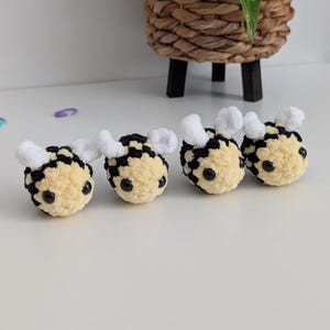 May include: Four handmade crochet bee ornaments. Each bee has a yellow body with black patches, black eyes, and white fuzzy wings. The bees are arranged in a row on a white surface. A woven basket with black legs is in the background.