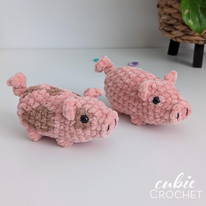 May include: Two handmade crochet pigs crafted from soft, plush pink yarn. One pig features a light brown patch on its side, while both have small curled tails, pointed ears, and black bead eyes. The text cubie CROCHET appears in the bottom right corner.