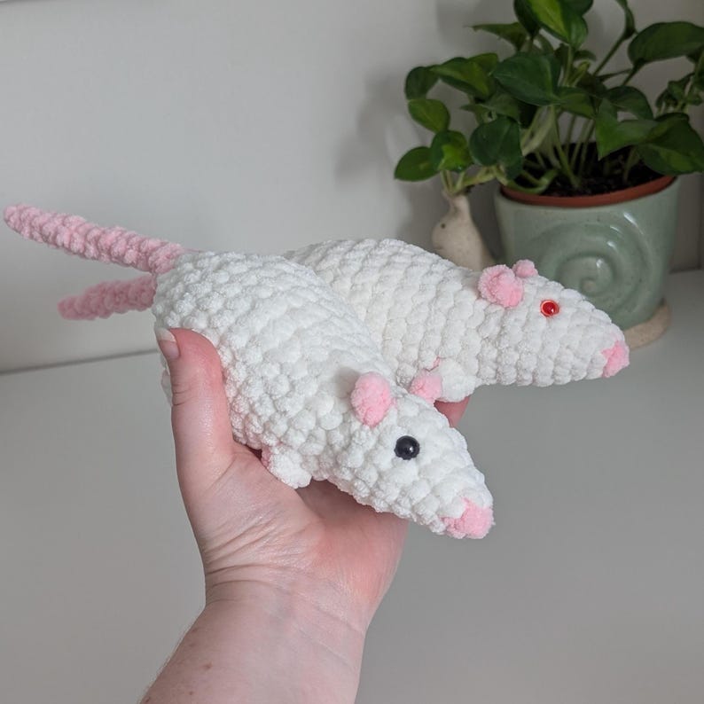 A hand holding two cream colored rat plushies. One has black eyes and the other has red eyes. They both have light pink noses, ears, and tails.