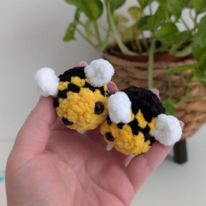 May include: Two handmade, plush bee toys. The bees are yellow and black, with white pom-pom wings and black bead eyes. The toys are held in a person's hand, with a blurred background of greenery.