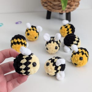 May include: A collection of handmade crochet bees in yellow, black, and white. The bees have small white wings and antennae, and black bead eyes. The bees are arranged on a white surface.