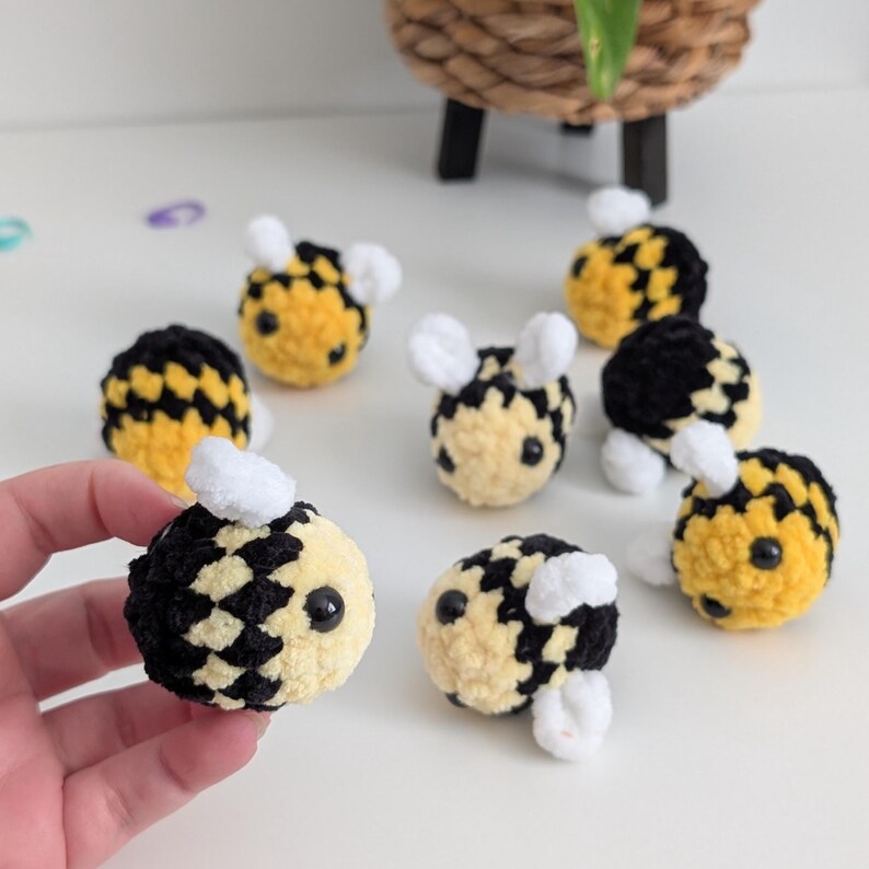 May include: A collection of handmade crochet bees in yellow, black, and white. The bees have small white wings and antennae, and black bead eyes. The bees are arranged on a white surface.