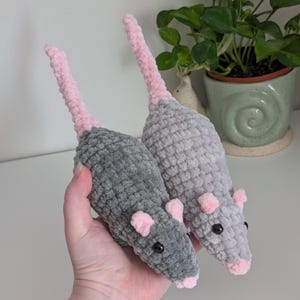 A hand holding up two rat plushies. The one on the left is dark gray, and the one on the right is light gray. They both have light pink noses, ears, and tails.