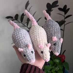 A hand holding up three crochet rat plushies, with their noses facing downwards and tails facing upwards. The one on the left is light gray, the one in the middle is a warm off-white color, and the one on the right is dark gray. They all have black eyes and light pink noses, ears, and tails.