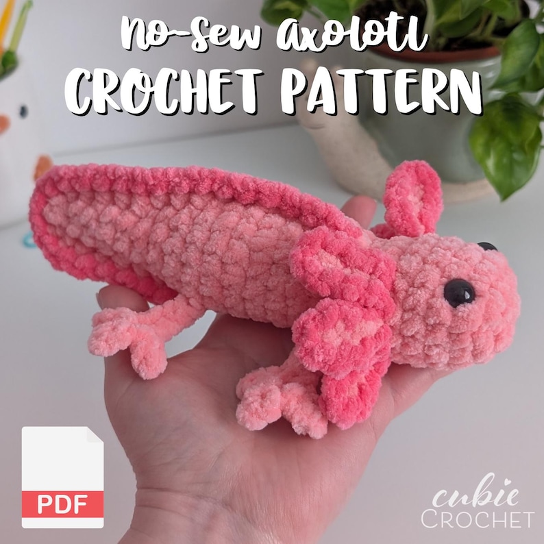 May include: A plush, pink axolotl crochet pattern. The handmade axolotl is a light and dark pink color with black eyes. The image includes the text "No-sew axolotl CROCHET PATTERN" and a PDF icon.