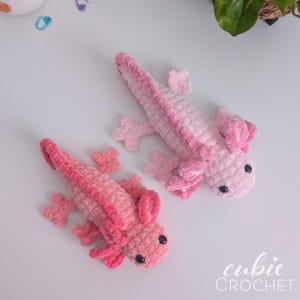 May include: Two handmade crochet axolotls in shades of pink. The axolotls have small black eyes and detailed gills. The image includes the text "cubic CROCHET" in the bottom right corner. These plush toys are perfect for a child's room.