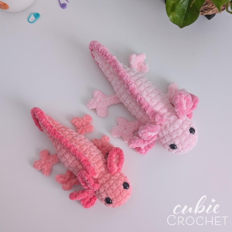 May include: Two handmade crochet axolotls in shades of pink. The axolotls have small black eyes and detailed gills. The image includes the text "cubic CROCHET" in the bottom right corner. These plush toys are perfect for a child's room.