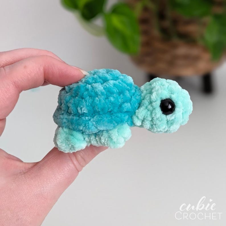 May include: A small, plush, crocheted turtle toy featuring a vibrant turquoise shell and a lighter mint green head and flippers. The turtle has a soft, fuzzy texture and a single visible black plastic eye. The text cubie CROCHET is in the corner.