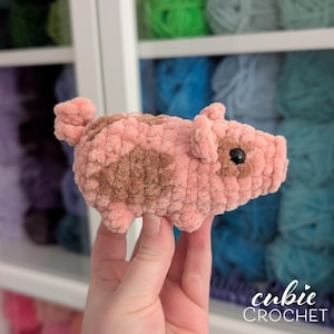 May include: A small, handmade crochet pig plushie crafted from soft, textured light pink yarn with brown patches on its side and face. The stuffed animal features a curled tail, two small ears, and a single visible black plastic bead eye.