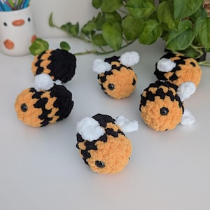 May include: A collection of six handmade crochet bees. Each bee features a fuzzy orange body with black markings, white wings, and black eyes. The bees are arranged on a white surface, with greenery in the background.