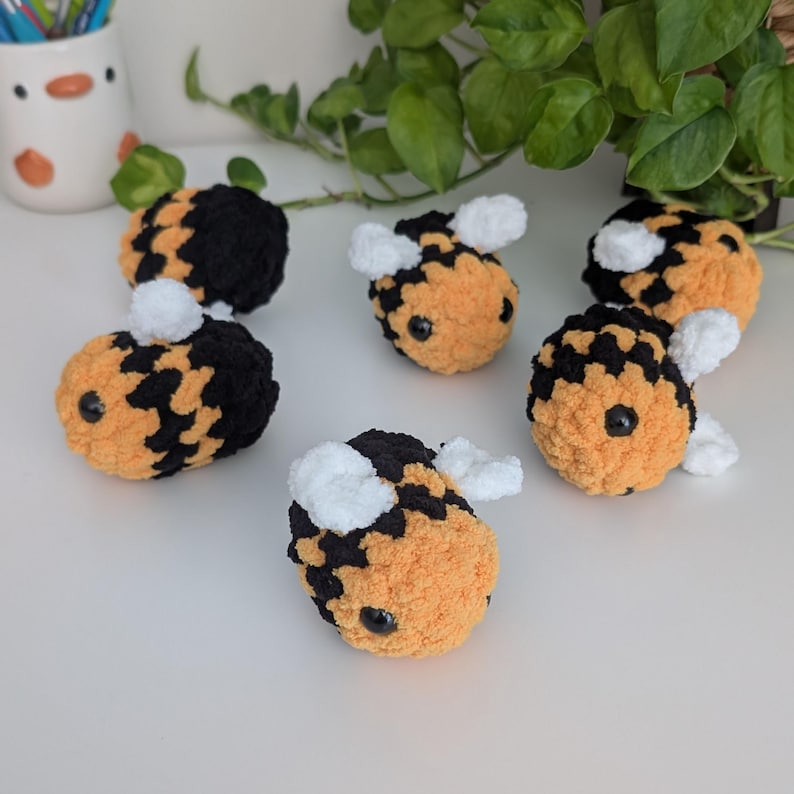 May include: A collection of six handmade crochet bees. Each bee features a fuzzy orange body with black markings, white wings, and black eyes. The bees are arranged on a white surface, with greenery in the background.