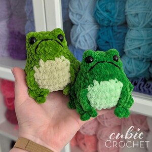 Op de afbeelding: Twee pluche kikker-speeltjes gemaakt van groen garen met zwarte plastic ogen. De grotere kikker heeft een cr&egrave;mekleurige buik, terwijl de kleinere kikker een lichtgroene buik heeft. Beide kikkers hebben een gestikte mond en een zachte textuur.
