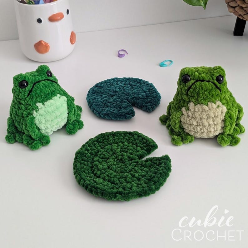 Op de afbeelding: Twee gehaakte pluche kikkers in groentinten met lichtgroene buiken en zwarte ogen, vergezeld door twee donkergroene ronde gehaakte waterleliebladeren. De tekst cubie CROCHET is zichtbaar in de rechterbenedenhoek van de afbeelding.