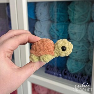 May include: A small, handmade crochet turtle plushie featuring a soft, textured light green body and a rounded, fuzzy peach-colored shell. The turtle has a single visible black plastic safety eye on its side, showcasing a compact, plush design.