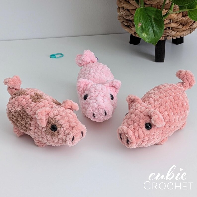 May include: Three handmade crochet pigs crafted from soft, plush pink yarn. Each pig features a curled tail, small pointed ears, and black bead eyes. One pig has brown patches on its back, while the others are solid pink. The text cubie CROCHET is visible.