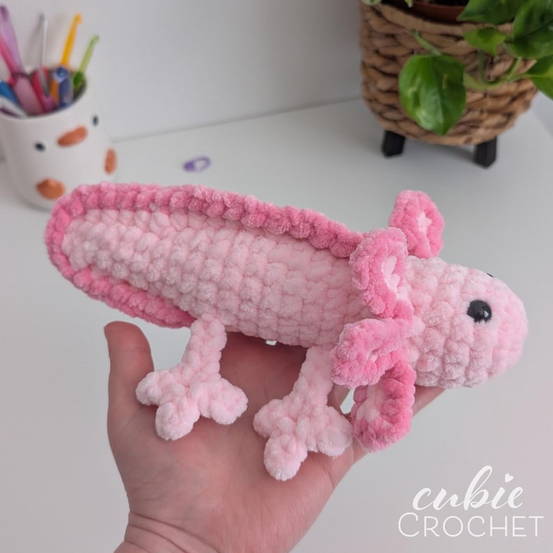 May include: A handmade, plush axolotl in shades of pink. The crocheted creature features a textured body, detailed gills, and small, black bead eyes. The axolotl is a soft, cuddly toy.