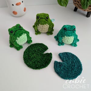 Op de afbeelding: Drie kleine, gehaakte pluche kikkers in verschillende tinten groen met zwarte kraaloogjes en lichtgroene buikjes, vergezeld door twee donkergroene gehaakte leliebladeren. De tekst "cubie CROCHET" is zichtbaar in de rechterbenedenhoek.