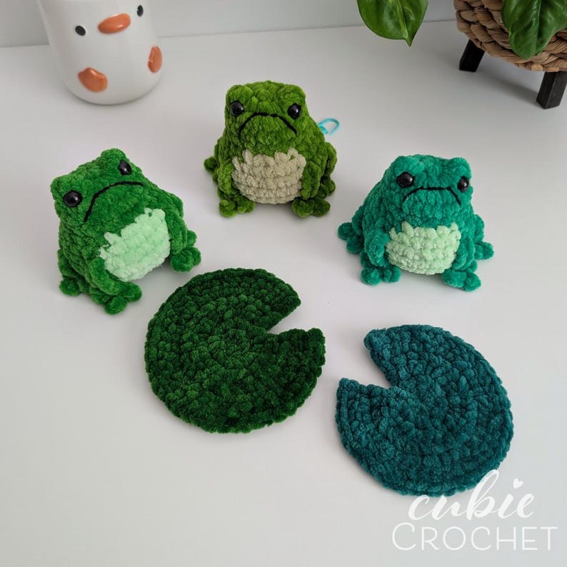 Op de afbeelding: Drie kleine, gehaakte pluche kikkers in verschillende tinten groen met zwarte kraaloogjes en lichtgroene buikjes, vergezeld door twee donkergroene gehaakte leliebladeren. De tekst "cubie CROCHET" is zichtbaar in de rechterbenedenhoek.