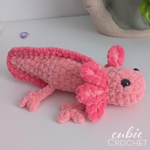 May include: A handmade, pink crochet axolotl plush toy. The plush features a textured body, detailed gills, and small black eyes. The axolotl is lying on a white surface. The text "cubie CROCHET" is in the bottom right corner.