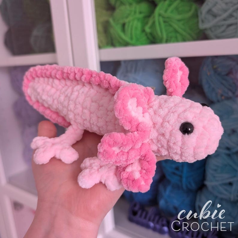 May include: A handmade pink and white crochet axolotl plush toy. The axolotl has a textured body, detailed gills, and small black eyes. The plush is held in a hand, with a background of colorful yarn.