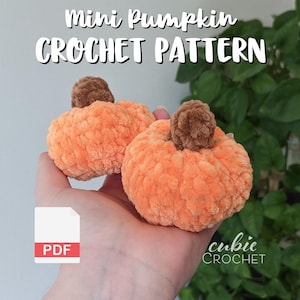 May include: Two orange crochet pumpkins with brown stems are held in a hand. The pumpkins have a textured, plush appearance. Text at the top reads "Mini Pumpkin Crochet Pattern". A PDF icon is in the lower left corner, and the "cubic CROCHET" logo is in the lower right.