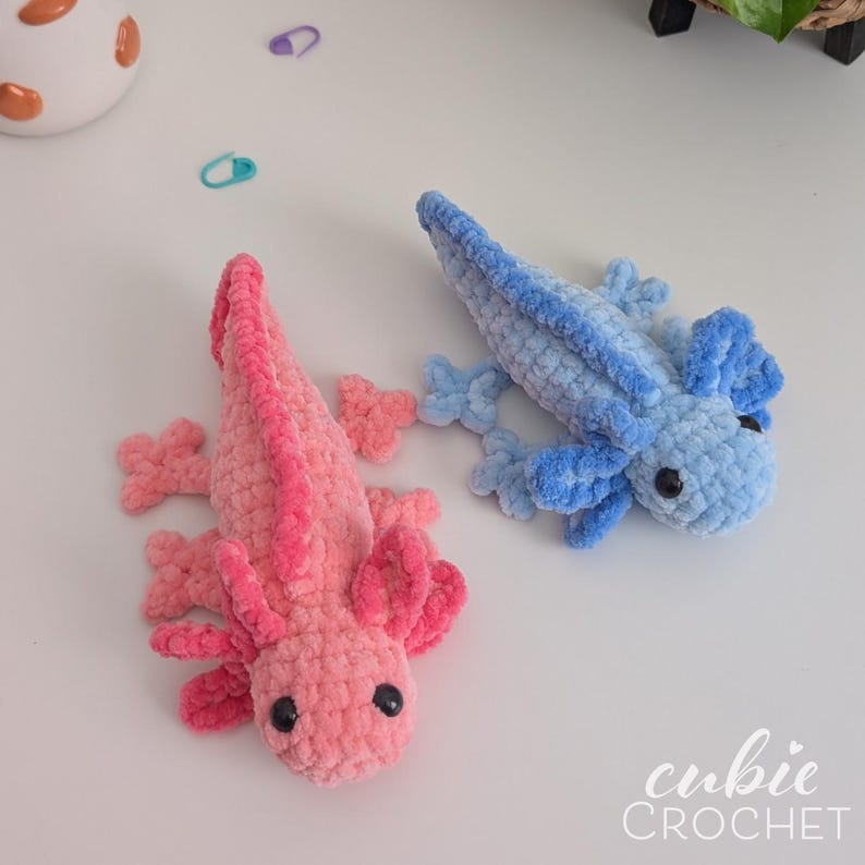 May include: Two handmade crochet axolotls, one pink and one blue, are displayed on a white surface. The pink axolotl has a darker pink frill and the blue axolotl has a lighter blue frill. Both have black eyes. The text "cubie CROCHET" is in the bottom right corner.