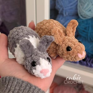 Op de afbeelding: Twee handgemaakte gehaakte konijntjes. E&eacute;n is grijs en wit, de andere is bruin. Beide hebben zwarte ogen en roze neuzen. De konijntjes zijn klein en zacht, perfect voor een kinderspeeltje of kinderkamerdecoratie. De tekst "cubie CROCHET" staat rechtsonder.