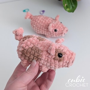 May include: Two small, handmade crochet pigs crafted from soft, plush pink yarn. Each pig features a curly tail, small ears, and dark, round plastic eyes. One pig has a distinct brown patch on its side and around its eye, while the other is solid pink.