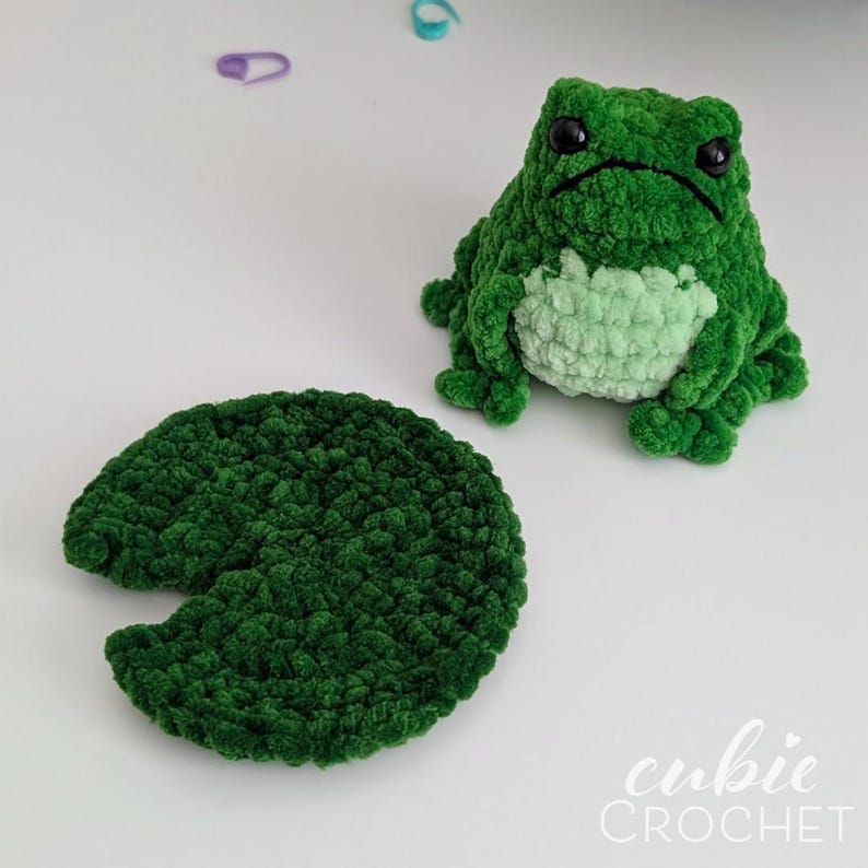 Op de afbeelding: Een donkergroene gehaakte kikkerknuffel met een lichtgroene buik en zwarte plastic ogen, vergezeld van een apart donkergroen gehaakt lelieblad. De tekst "cubie CROCHET" is zichtbaar in de rechterbenedenhoek van de afbeelding.