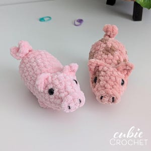 May include: Two handmade crochet plush pigs made from soft, textured yarn. One pig is solid light pink, while the other features a mottled pattern of light pink and brown. Both have small ears, curled tails, and black plastic eyes. Text reads cubie CROCHET.
