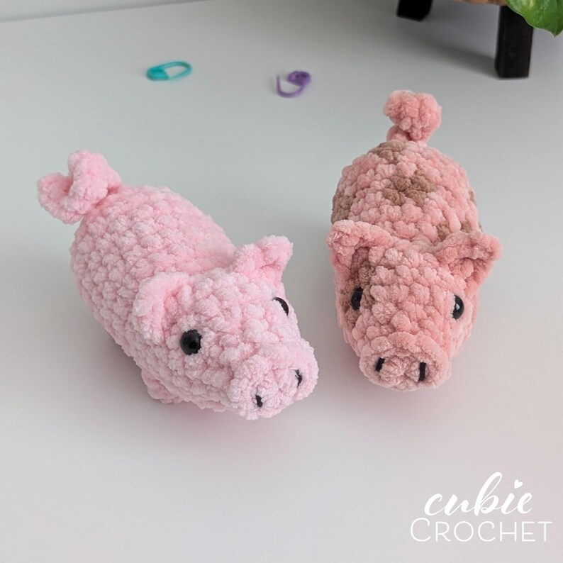 May include: Two handmade crochet plush pigs made from soft, textured yarn. One pig is solid light pink, while the other features a mottled pattern of light pink and brown. Both have small ears, curled tails, and black plastic eyes. Text reads cubie CROCHET.