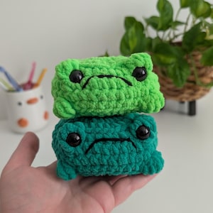May include: Two stacked crochet frog plushies made of soft yarn. The top frog is bright lime green, and the bottom frog is a darker forest green. Both feature large, round, black plastic safety eyes and a simple black stitched mouth line.