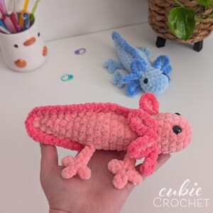 May include: A handmade, plush axolotl in shades of pink, with a darker pink outline. A smaller blue axolotl is in the background. The axolotls have small black eyes and detailed gills. The text "cubie CROCHET" is in the bottom right corner.