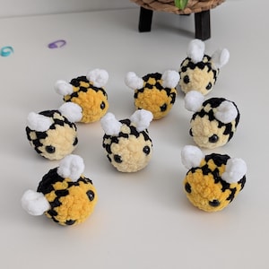 May include: A collection of handmade crochet bees. Each bee features a yellow body with black stripes, white fuzzy wings, and small black eyes. The bees are arranged on a white surface, showcasing their detailed craftsmanship.