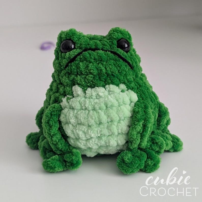 Op de afbeelding: Een pluche kikker gemaakt van donkergroen garen met een licht mintgroene buik. De kikker heeft twee grote, ronde, zwarte plastic ogen en een gestikte mondlijn. De tekst "cubie CROCHET" staat in het wit rechtsonder afgedrukt.