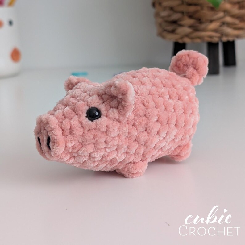 May include: A small, pink, plush pig made of soft, textured yarn. The pig features two small ears, a snout with two black stitched nostrils, and two round, black plastic eyes. A small, curled tail is visible on the back. The text cubie CROCHET is present.