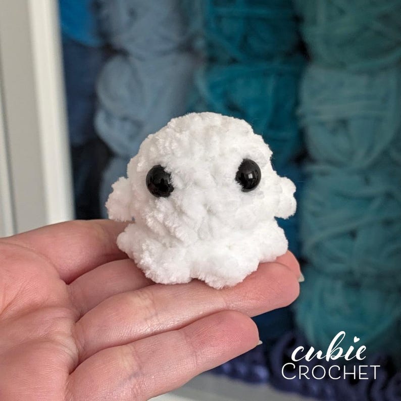 K&ouml;nnte beinhalten: Ein kleines, wei&szlig;es, geh&auml;keltes Pl&uuml;schwesen mit rundem K&ouml;rper und zwei auff&auml;lligen, glatten, schwarzen Kunststoffaugen. Die Textur des Garns ist weich und flauschig, was eine kompakte, abgerundete Form mit kleinen seitlichen Gliedma&szlig;en ergibt.