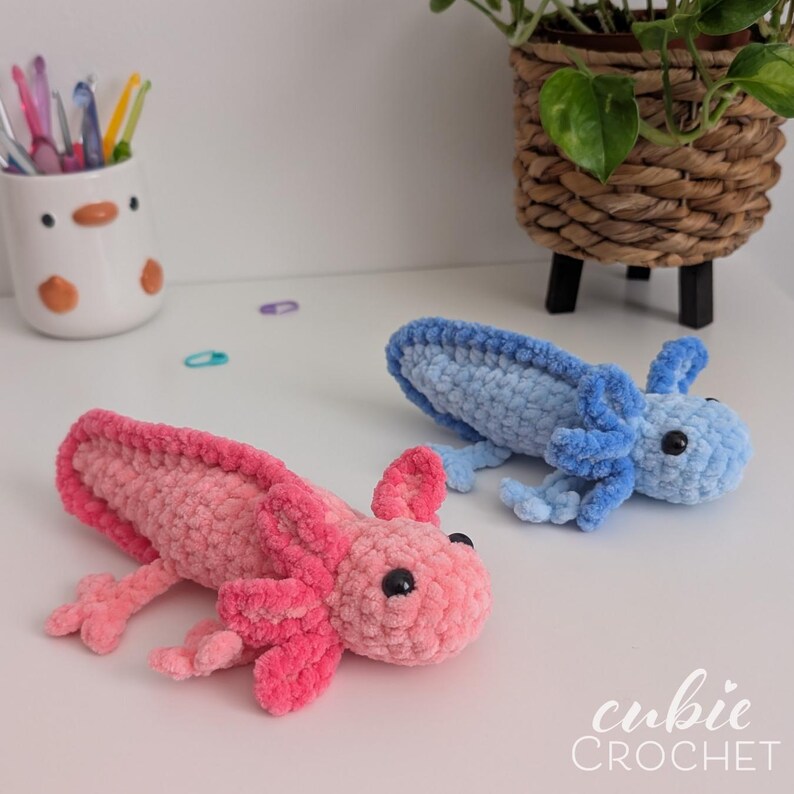 May include: Two handmade crochet axolotls in the image. One is pink, the other is blue. The axolotls have small black eyes and detailed gills. The axolotls are on a white surface. The image also includes a pen holder and a potted plant.