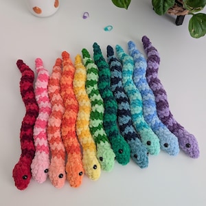 May include: A collection of eleven plush snakes made from soft, textured yarn, arranged in a row. Each snake features a distinct color, including red, pink, coral, orange, yellow, light green, dark green, navy, teal, light blue, and purple.