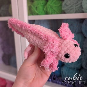 May include: A handmade pink crochet axolotl with black safety eyes. The axolotl has fluffy, textured gills and a soft, plush body. The image includes the text "cubie CROCHET".