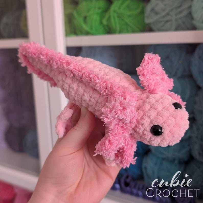 May include: A handmade pink crochet axolotl with black safety eyes. The axolotl has fluffy, textured gills and a soft, plush body. The image includes the text "cubie CROCHET".