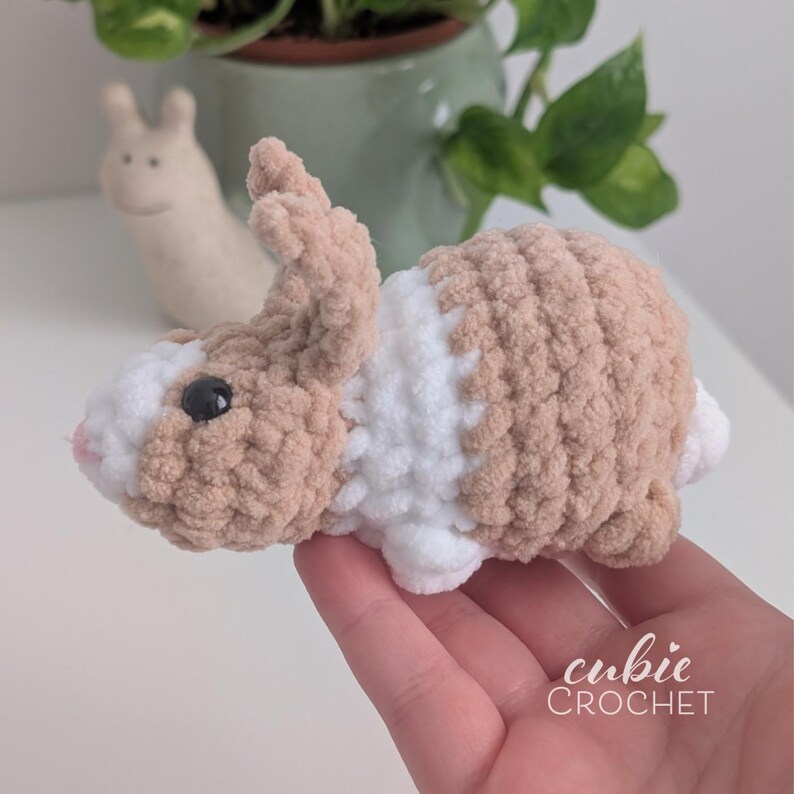 Op de afbeelding: Een handgemaakt gehaakt konijn in tinten van bruin, wit en zwart. Het konijn heeft een zachte textuur en een roze neus. De tekst "cubic CROCHET" is zichtbaar. Een schattig, zacht speelgoed.