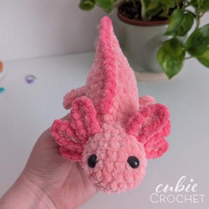 May include: A handmade pink crochet axolotl plush toy with black safety eyes. The axolotl has a textured, plush appearance and features detailed gills and a dorsal fin. The text "cubic CROCHET" is in the bottom right corner.
