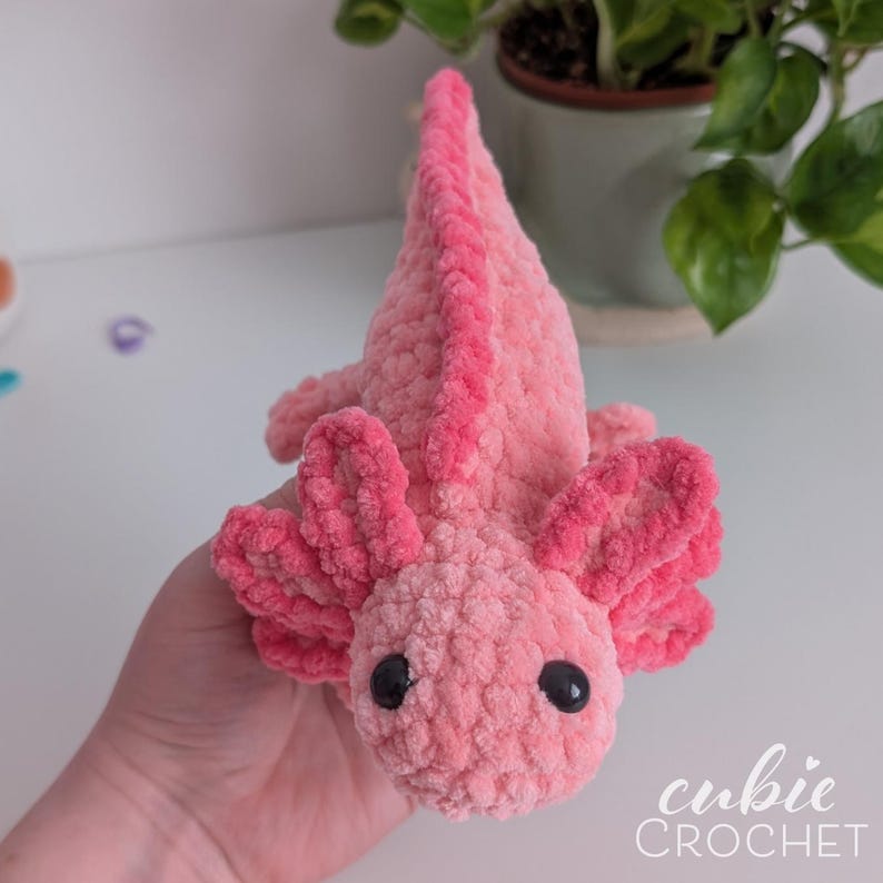 May include: A handmade pink crochet axolotl plush toy with black safety eyes. The axolotl has a textured, plush appearance and features detailed gills and a dorsal fin. The text "cubic CROCHET" is in the bottom right corner.