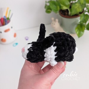 Op de afbeelding: Een handgemaakte gehaakte konijn in zwart en wit. Het konijn heeft een zachte textuur, met zwarte oren en een witte buik. De neus van het konijn is roze. De tekst "cubic CROCHET" is zichtbaar in de rechter benedenhoek. Het konijn wordt in een hand gehouden.